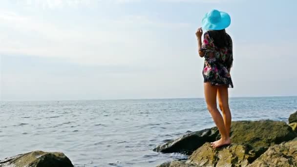 Mujer joven en un colorido vestido de verano admirando el mar en la costa rocosa — Vídeos de Stock