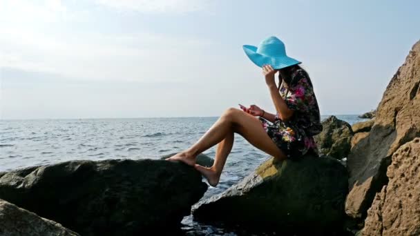 Mladá žena v letní šaty a modrý klobouk sedí na skalní mořského pobřeží, smartphone ve svých rukou