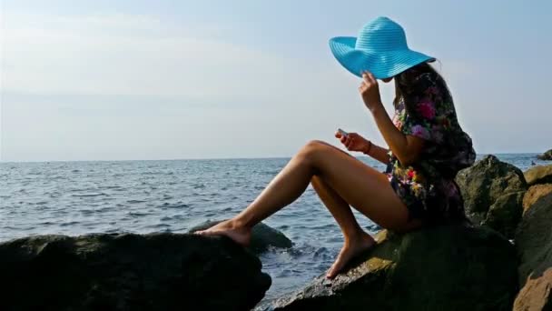 Молодая женщина в летнем платье и синей шляпе сидит на скалистом берегу моря, смартфон в руках — стоковое видео