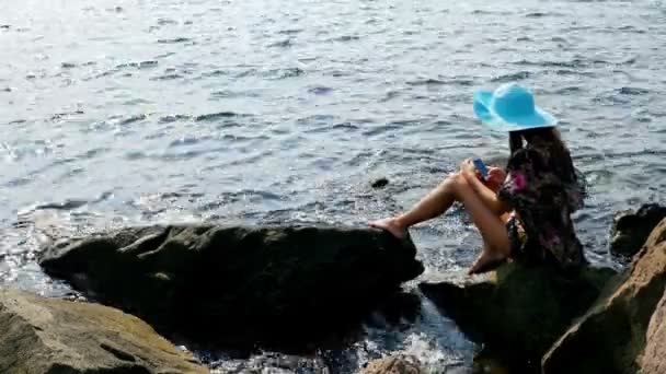 Mladá žena v letní šaty a modrý klobouk sedí na skalní mořského pobřeží, smartphone ve svých rukou — Stock video