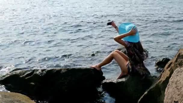 Mladá žena v letní šaty a modrý klobouk sedí na skalní mořského pobřeží, smartphone ve svých rukou — Stock video