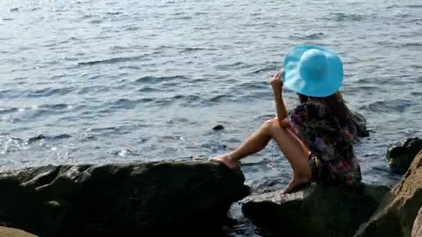 Siluet seorang wanita muda dengan gaun musim panas berwarna-warni dan topi biru di pantai laut berbatu — Stok Video