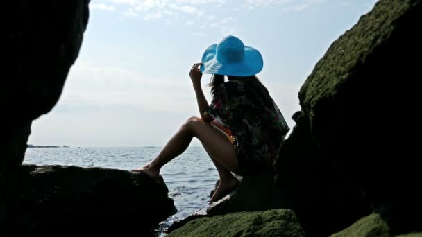 Silhuetten av en ung kvinna i en färgglad sommarklänning och blå hatt på rocky havet — Stockvideo