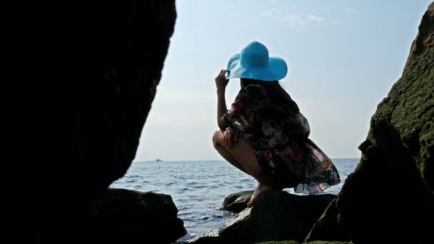 Silhouette of a young woman in a colorful summer dress and blue hat on the rocky sea shore — Stock Video