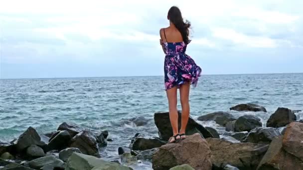 Mladá žena, sedící na skalnatém pobřeží u moře při západu slunce, její šaty vlající ve větru — Stock video