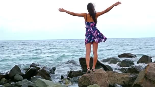 Jonge vrouw zittend op de rotsachtige kust door de zee bij zonsondergang, haar jurk wapperen in de wind — Stockvideo