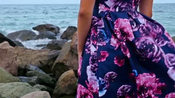 Junge Frau sitzt bei Sonnenuntergang am felsigen Ufer am Meer, ihr Kleid flattert im Wind — Stockvideo