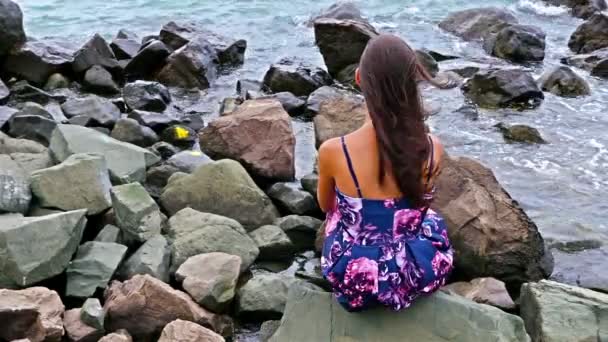 Junge Frau sitzt bei Sonnenuntergang am felsigen Ufer am Meer, ihr Kleid flattert im Wind — Stockvideo