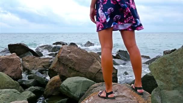 Молодая женщина, сидящая на скалистом берегу у моря на закате, ее платье развевается на ветру — стоковое видео