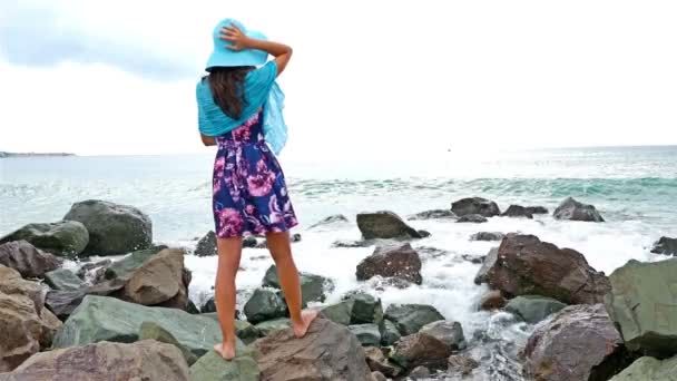 Young woman with summer dress, scarf and hat standing on the rocky shore by the sea at sunset, sea waves crash on the rocks — Stock Video