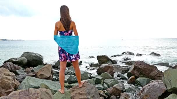 Jovem mulher com vestido de verão, cachecol e chapéu em pé na costa rochosa junto ao mar ao pôr do sol, ondas do mar bater sobre as rochas — Vídeo de Stock