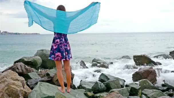 Junge Frau mit Sommerkleid, Schal und Mütze steht am felsigen Ufer am Meer bei Sonnenuntergang, Meereswellen krachen auf die Felsen — Stockvideo