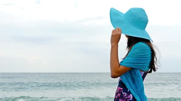 Junge Frau mit Sommerkleid, Schal und Mütze steht am felsigen Ufer am Meer bei Sonnenuntergang, Meereswellen krachen auf die Felsen — Stockvideo