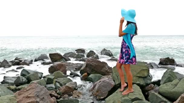 夏天的衣服、 围巾和帽子站在多岩石的海岸的海上日落，海浪撞碎在岩石上的年轻女子 — 图库视频影像