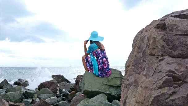 Mujer joven con vestido de verano, bufanda y sombrero de pie en la orilla rocosa junto al mar al atardecer, las olas del mar chocan contra las rocas — Vídeos de Stock