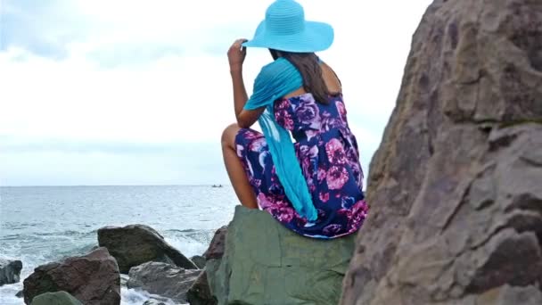 Mujer joven con vestido de verano, bufanda y sombrero sentado en la orilla rocosa junto al mar al atardecer, las olas del mar chocan contra las rocas, dolly — Vídeos de Stock