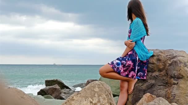 Mujer joven con vestido de verano, bufanda y sombrero de pie en la orilla rocosa junto al mar al atardecer, las olas del mar chocan contra las rocas — Vídeo de stock
