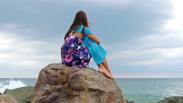 Jovem mulher com vestido de verão, cachecol e chapéu em pé na costa rochosa junto ao mar ao pôr do sol, ondas do mar bater sobre as rochas — Vídeo de Stock