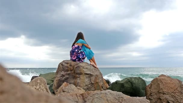 Ung kvinna med sommarklänning, scarf och hatt stående på den steniga stranden vid havet vid solnedgången, havet vågor krasch på klipporna — Stockvideo
