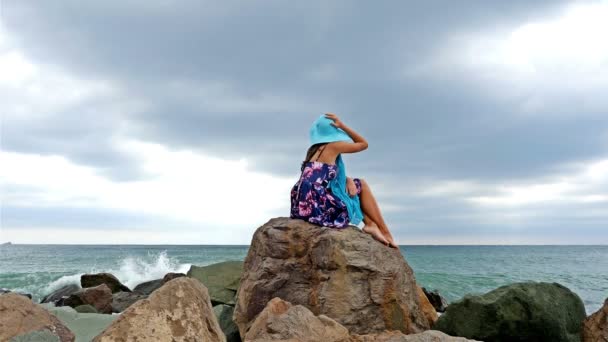 夏天的衣服、 围巾和帽子站在多岩石的海岸的海上日落，海浪撞碎在岩石上的年轻女子 — 图库视频影像