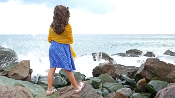 Liten flicka i en klänning och halsduk stående på klipporna, njuter av det stormande havet — Stockvideo