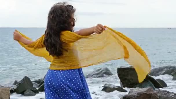 Menina em um vestido e cachecol em pé sobre as rochas e olhando para o mar tempestuoso — Vídeo de Stock