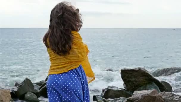 Petite fille en robe et écharpe debout sur les rochers et regardant vers la mer orageuse — Video