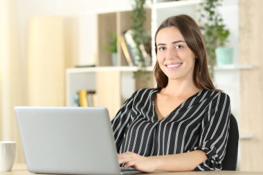 Evdeki masada dizüstü bilgisayar kullanarak kameraya bakan mutlu bir kadın.