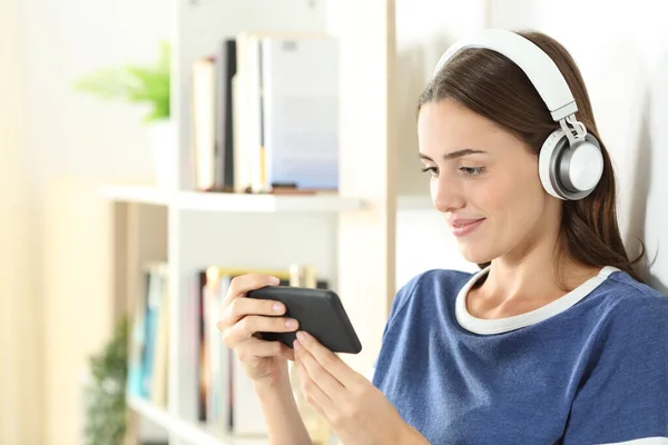 Uśmiechnięta Nastolatka Ogląda Media Smartfonie Nosząc Słuchawki Stojące Salonie Domu — Zdjęcie stockowe