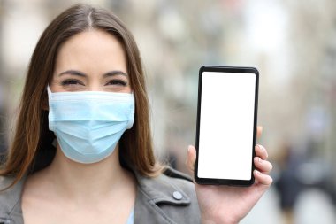 Front view portrait of a happy woman wearing mask to avoid coronavirus or pollution showing blank smart phone screen mock up clipart