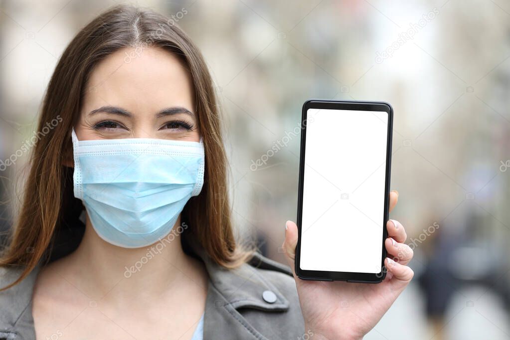 Front view portrait of a happy woman wearing mask to avoid coronavirus or pollution showing blank smart phone screen mock up