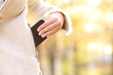 Close up of woman hand keeping smart phone in a jacket pocket in fall season in a park clipart