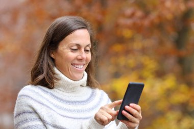 Mutlu yetişkin kadın, sonbahar mevsiminde parkta akıllı telefon kullanıyor.