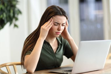 Stressed woman suffering migraine with a laptop sitting in a table at home clipart