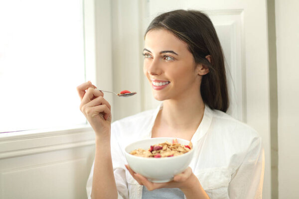 Happy woman eating cereals at breakfast in the morning looking through a window at home
