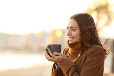 Kışın kahve içen mutlu kadın gün batımında sahile bakıyor.