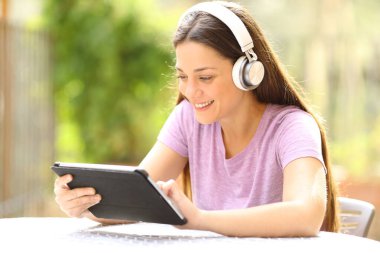 Kablosuz kulaklıklı mutlu kadın evdeki bir bahçede tablet içeriğini kontrol ediyor.