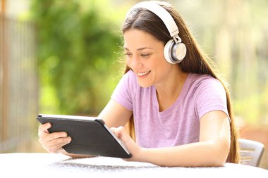 Kablosuz kulaklıklı mutlu kadın evdeki bir bahçede tablet içeriğini kontrol ediyor.
