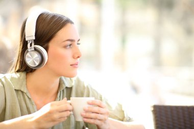 Kablosuz kulaklık takan, müzik dinleyen kahve dükkanında kupa tutan düşünceli bir kadın.