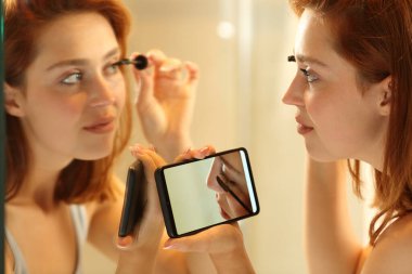 Attentive woman putting on make up watching video tutorial looking at mirror in the bathroom at home