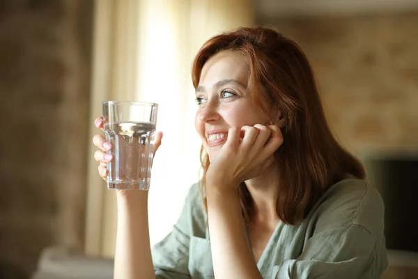 快乐的女人举着水杯坐在客厅里看着远方 — 图库照片