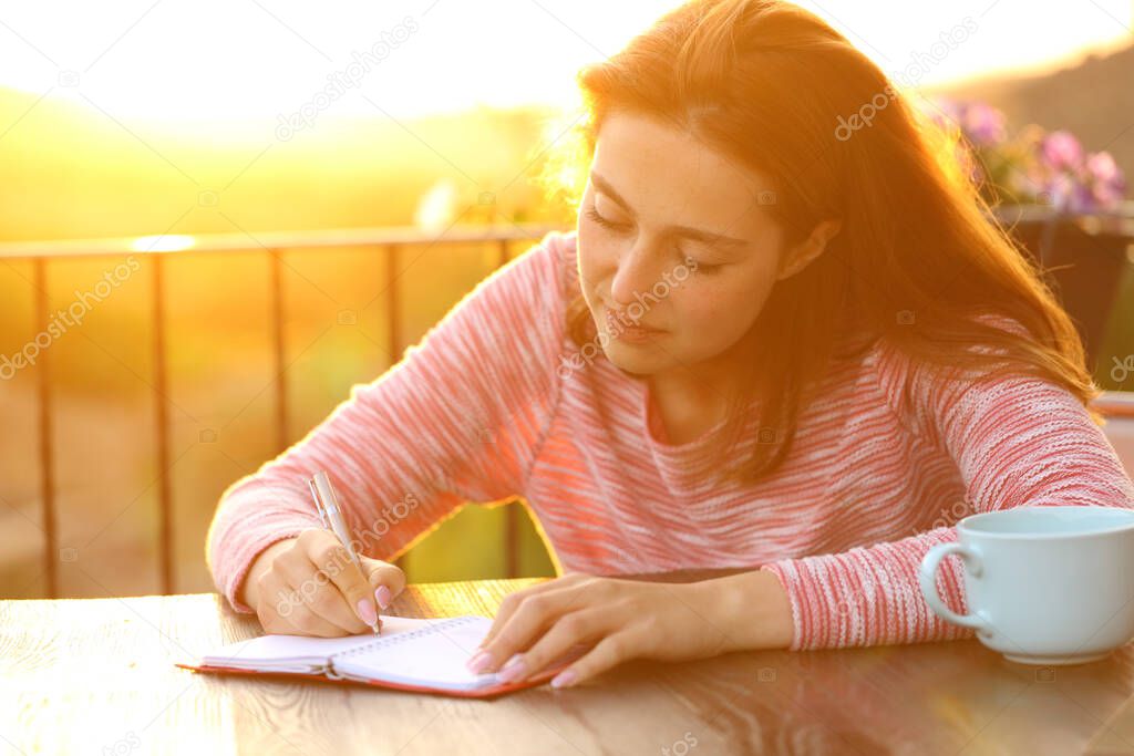 Concentrated woman writing on paper agenda at sunset in a balcony
