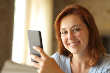 Happy woman holding smart phone looks at camera sitting at home