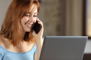 Happy woman talking on mobile phone using laptop in the bedroom at home