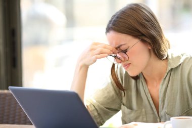 Tired woman wearing eyeglasses suffering eyestrain using laptop sitting in a bar