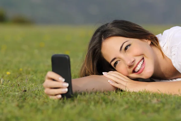 Mooi meisje lachend met behulp van een slimme telefoon liggen op het gras — Stockfoto