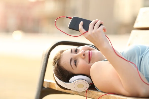 Teen flicka lyssnar musik från en telefon liggande i en bänk — Stockfoto