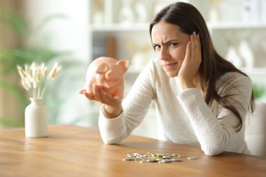 Öfkeli zavallı kadın, evdeki ahşap bir masada birkaç birikimi kontrol edip şikâyet ediyor.