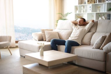 Evdeki rahat bir kanepede oturan adam büyük bir pencerenin yanında dinleniyor.