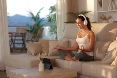 Mutlu yogi kulaklık takıyor dijital tablette yoga dersini izliyor gün batımında evdeki kanepede oturuyor.